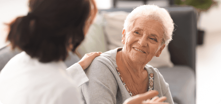 Support worker talking with older woman sitting on a couch, with her hand placed on the older woman's shoulder